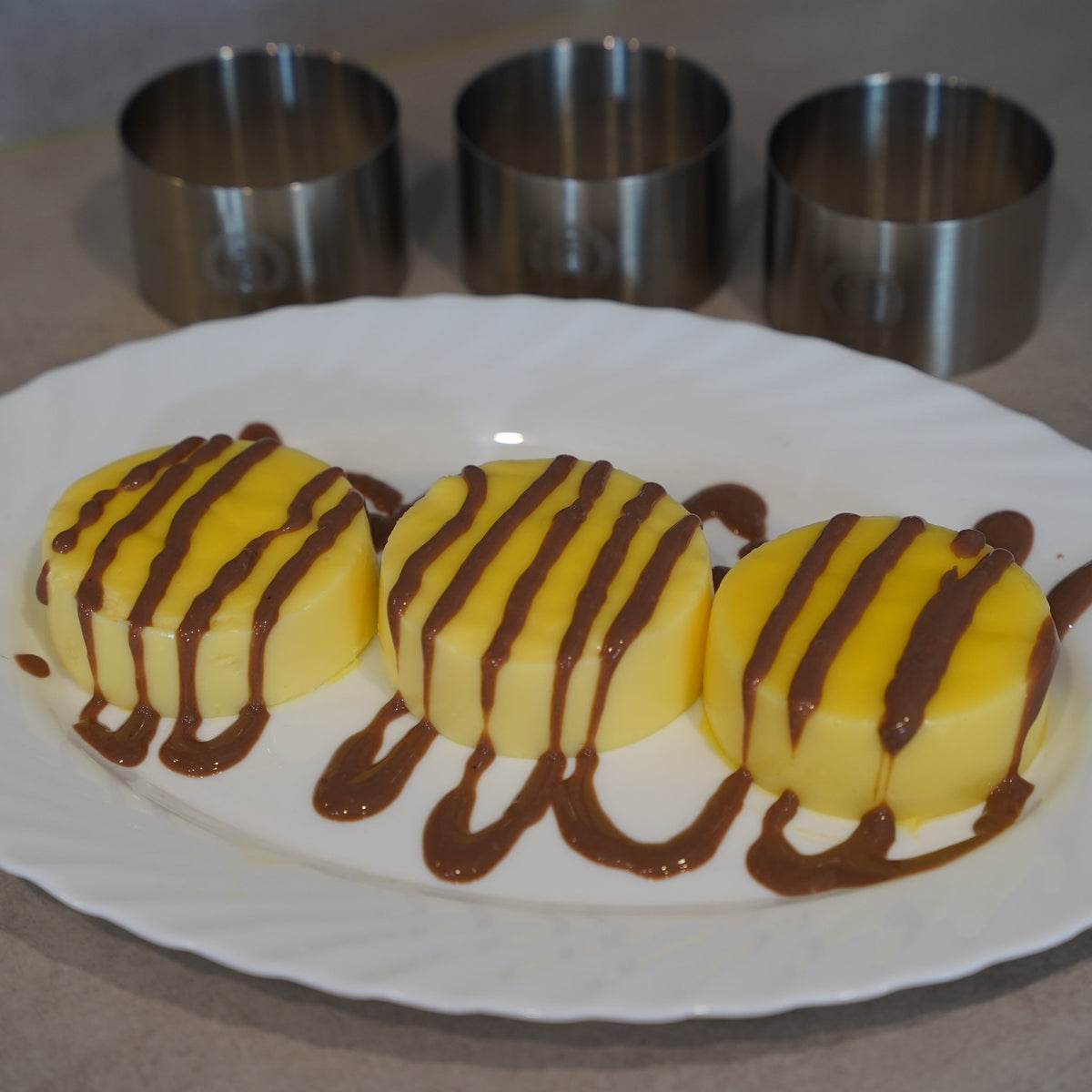 Drei Vanilledesserts aus dem Speisering mit Schokoladensauce, dahinter die Ringe